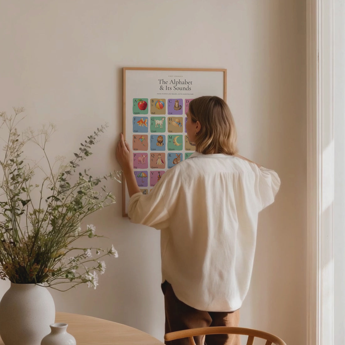 Watercolour alphabet poster in a natural wood frame, shown being placed on a wall followed by close and wide room views.